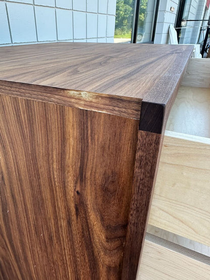 A close-up of the 3 Drawer Dresser in Walnut - SECOND by Krovel Furniture Co. highlights its dark-finished solid hardwood top and sides with natural wood grain patterns, while an open, unfinished drawer is visible; white tiles and a window appear in the background.