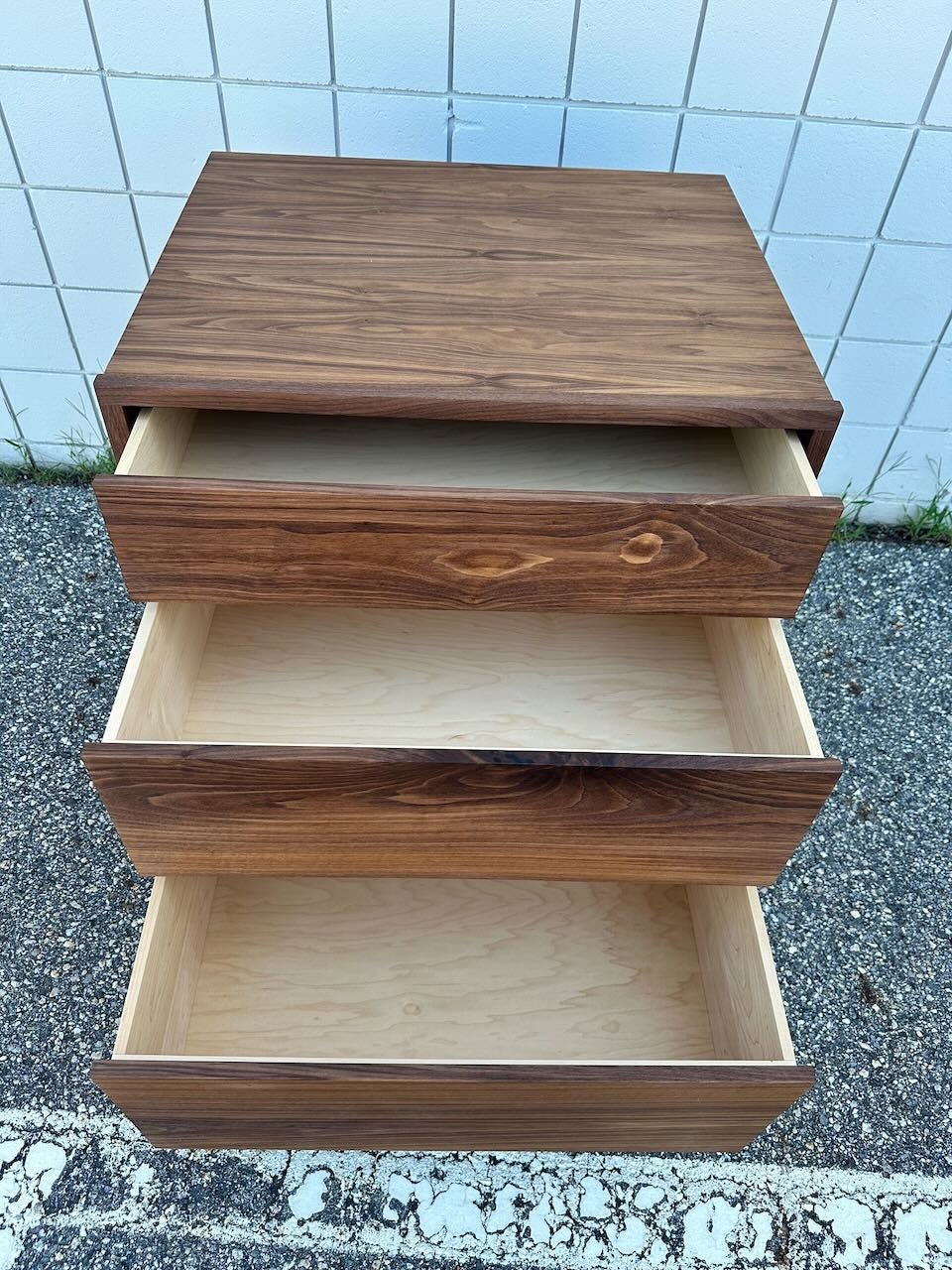 A solid hardwood 3 Drawer Dresser in Walnut - SECOND by Krovel Furniture Co., with the top two drawers slightly open, stands on outdoor pavement against a white tiled wall and features a zero-VOC finish for an eco-friendly touch.