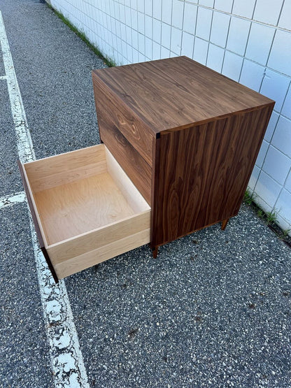 A Krovel Furniture Co. 3 Drawer Dresser in Walnut—with a dark, zero-VOC finish on solid hardwood—stands on asphalt beside a white tiled wall. One of its light-colored drawers is open, revealing the interior.