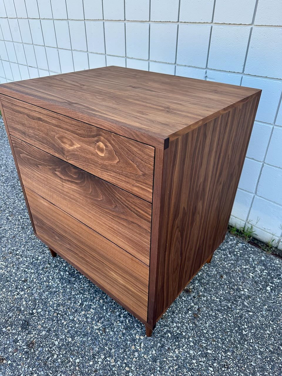 The Krovel Furniture Co. 3 Drawer Dresser in Walnut - SECOND sits on a gravel surface in front of a white tiled wall, its visible wood grain highlighted by a zero-VOC finish that enhances the dresser’s natural, modern appeal.