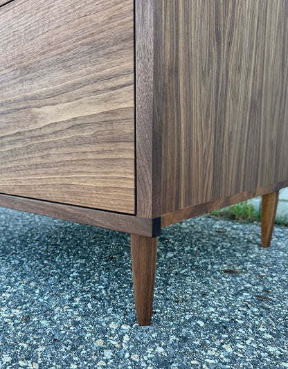 Close-up of the lower corner of the 3 Drawer Dresser in Walnut - SECOND by Krovel Furniture Co., showcasing its angled hardwood legs set on a textured outdoor surface. The walnut wood grain and cabinet details are accentuated by a zero-VOC finish.