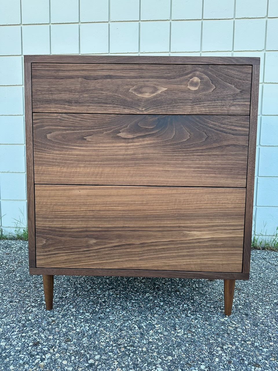 The Krovel Furniture Co. 3 Drawer Dresser in Walnut – SECOND is a mid-century modern piece crafted from solid hardwood with a visible wood grain, three spacious drawers, and four tapered legs. Finished with a zero-VOC coating, the dresser is shown outdoors on a textured surface against a tiled wall.