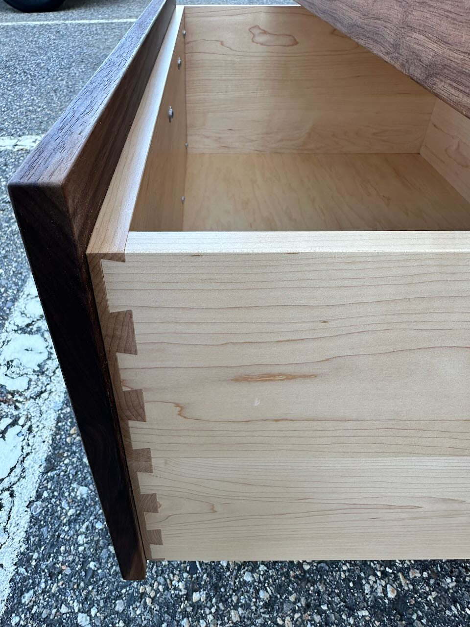 An open drawer from the 3 Drawer Walnut Dresser - SAMPLE by Krovel Furniture Co. showcases visible dovetail joints as it rests on a textured outdoor surface. The drawer front is finished in a rich walnut veneer, while the sides and interior are crafted from light maple wood—highlighting classic dovetailed maple construction and completed with an eco-friendly zero-VOC oil finish.