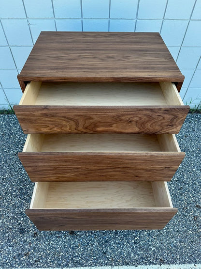 A 3 Drawer Walnut Dresser - SAMPLE by Krovel Furniture Co., featuring dovetailed maple drawers—all open—sits on pavement in front of a white tiled wall.