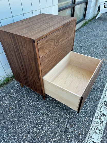 A 3 Drawer Walnut Dresser by Krovel Furniture Co. sits on a gravel surface near a white brick building and windows, with its bottom drawer open to reveal dovetailed maple drawers.