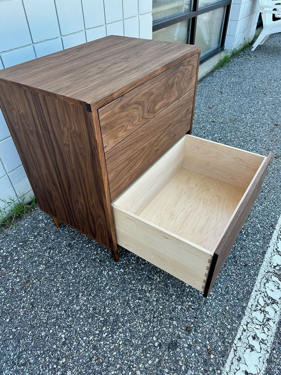 A 3 Drawer Walnut Dresser by Krovel Furniture Co. sits on a gravel surface near a white brick building and windows, with its bottom drawer open to reveal dovetailed maple drawers.