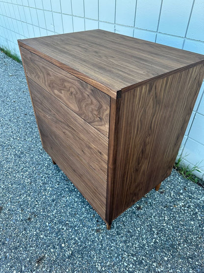 A 3 Drawer Walnut Dresser by Krovel Furniture Co., finished with a smooth, natural zero-VOC oil, stands on a textured outdoor surface beside a white tiled wall. The dresser’s clean lines and craftsmanship are highlighted by two visible dovetailed maple drawers.