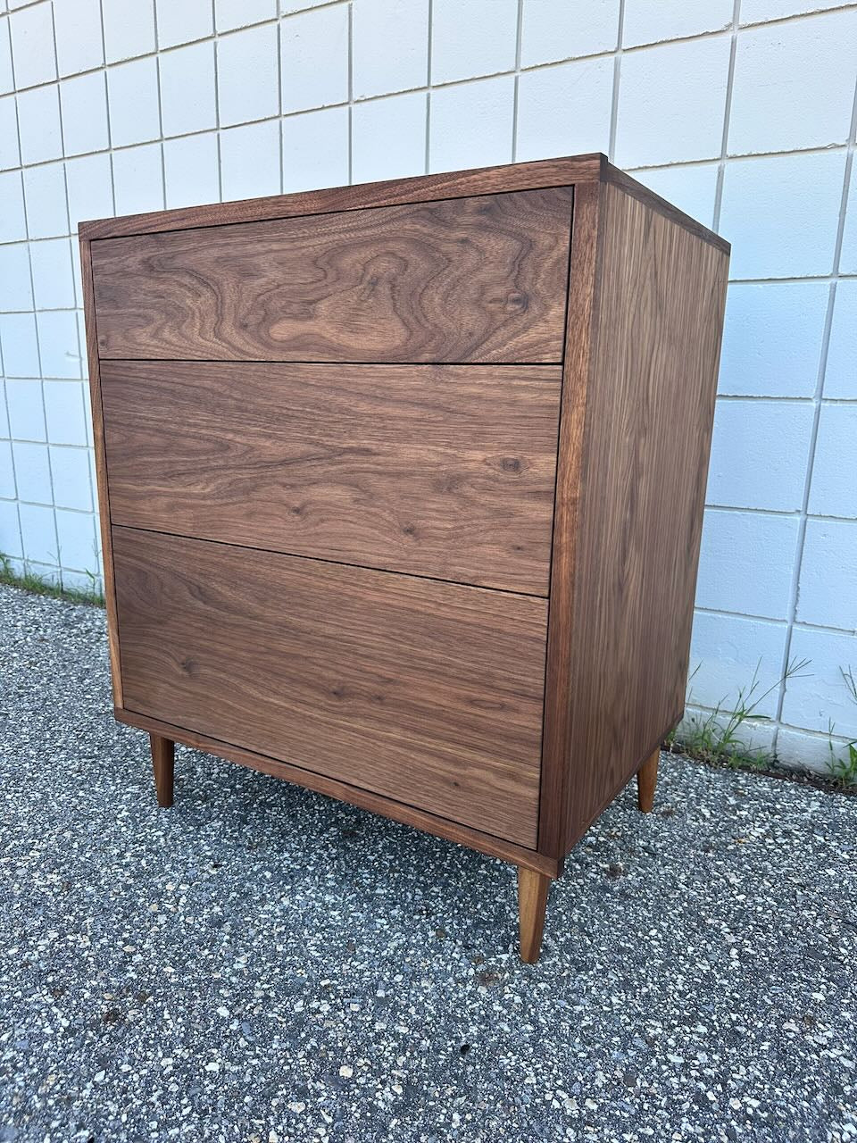 The 3 Drawer Walnut Dresser - SAMPLE by Krovel Furniture Co. showcases mid-century modern style with its walnut veneer construction, three dovetailed maple drawers, and tapered legs, seen here outdoors against a white tiled wall and gravel ground.