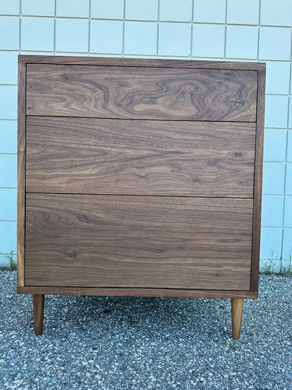 The 3 Drawer Walnut Dresser - SAMPLE by Krovel Furniture Co., featuring dovetailed maple drawers and a natural zero-VOC oil finish, stands on a gravel surface in front of a light-colored tiled wall.