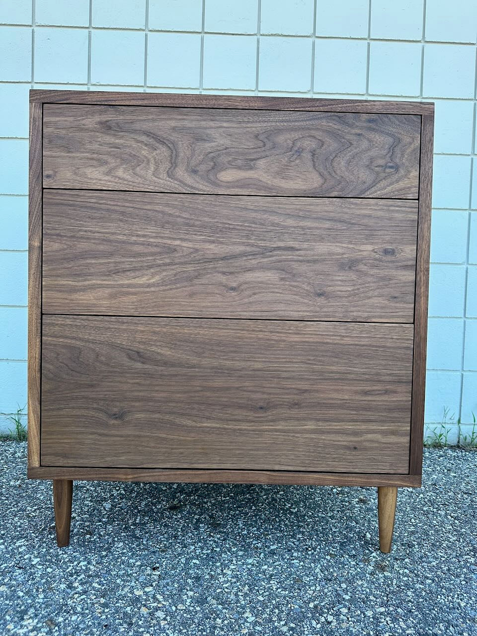 The 3 Drawer Walnut Dresser - SAMPLE by Krovel Furniture Co., featuring dovetailed maple drawers and a natural zero-VOC oil finish, stands on a gravel surface in front of a light-colored tiled wall.