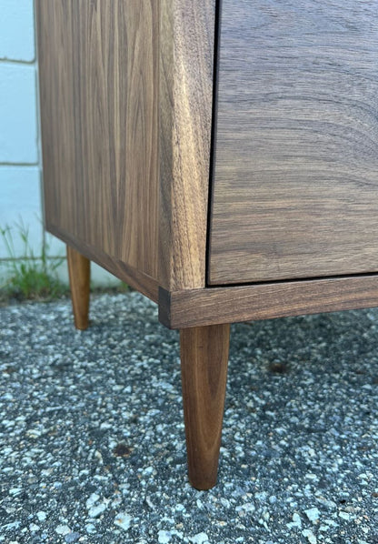 Close-up of the corner of the Krovel Furniture Co. 3 Drawer Walnut Dresser - SAMPLE, featuring tapered legs and standing on a rough, textured outdoor surface near a white brick wall with some grass growing at the edge.