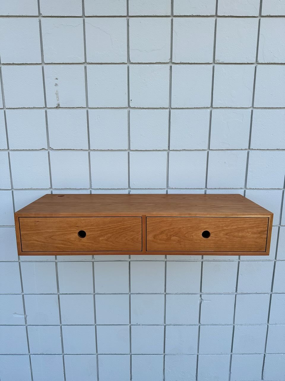 A Cherry Desk 40″ – SAMPLE by Krovel Furniture Co., featuring two solid Maple drawers with round hole handles, mounted on a white tiled wall.