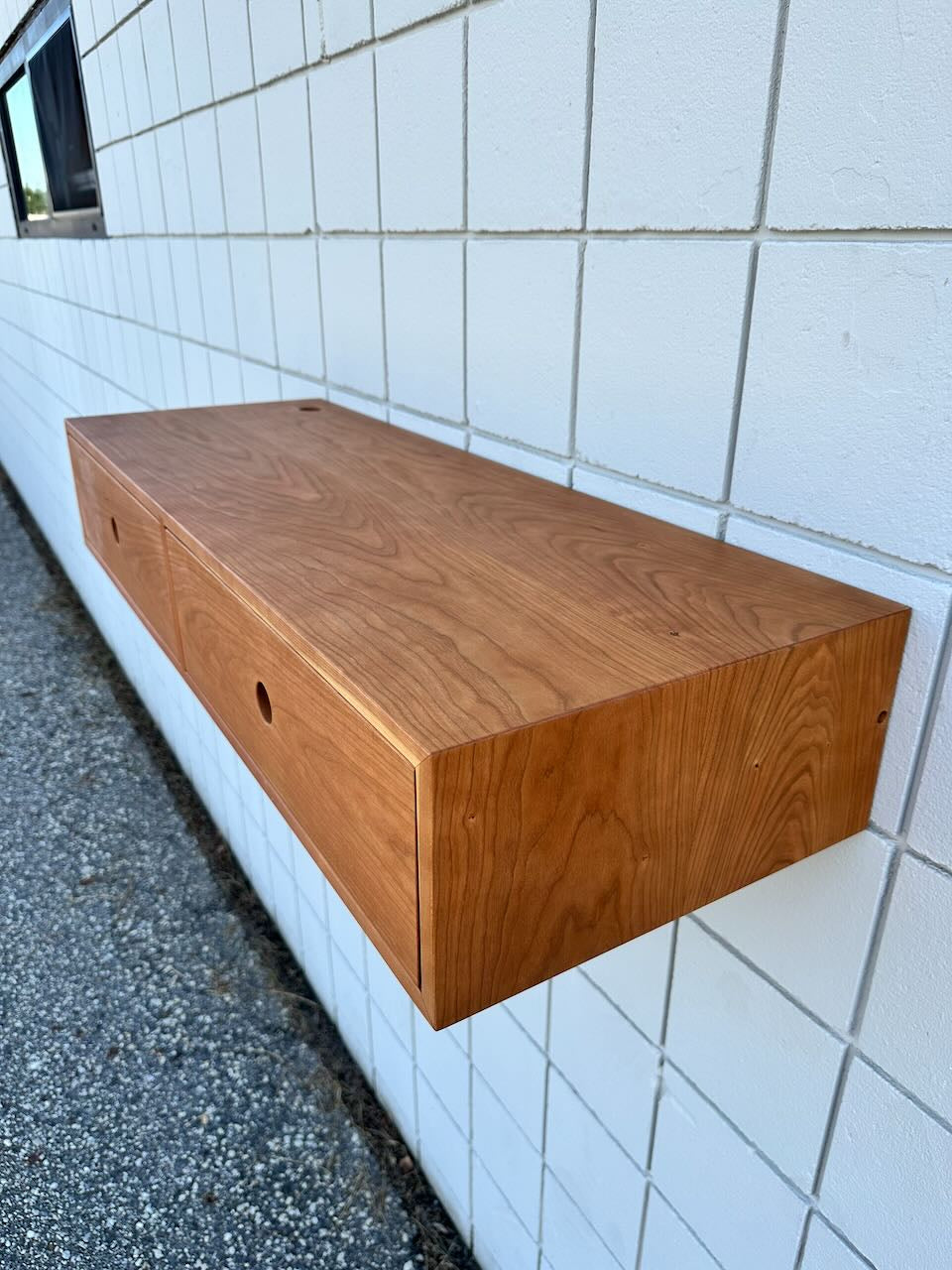 A Cherry Desk 40″ by Krovel Furniture Co. is mounted on a white tiled wall above a gravel surface, reflecting a minimalist design. The piece features solid cherry craftsmanship, combining elegant construction with modern simplicity.