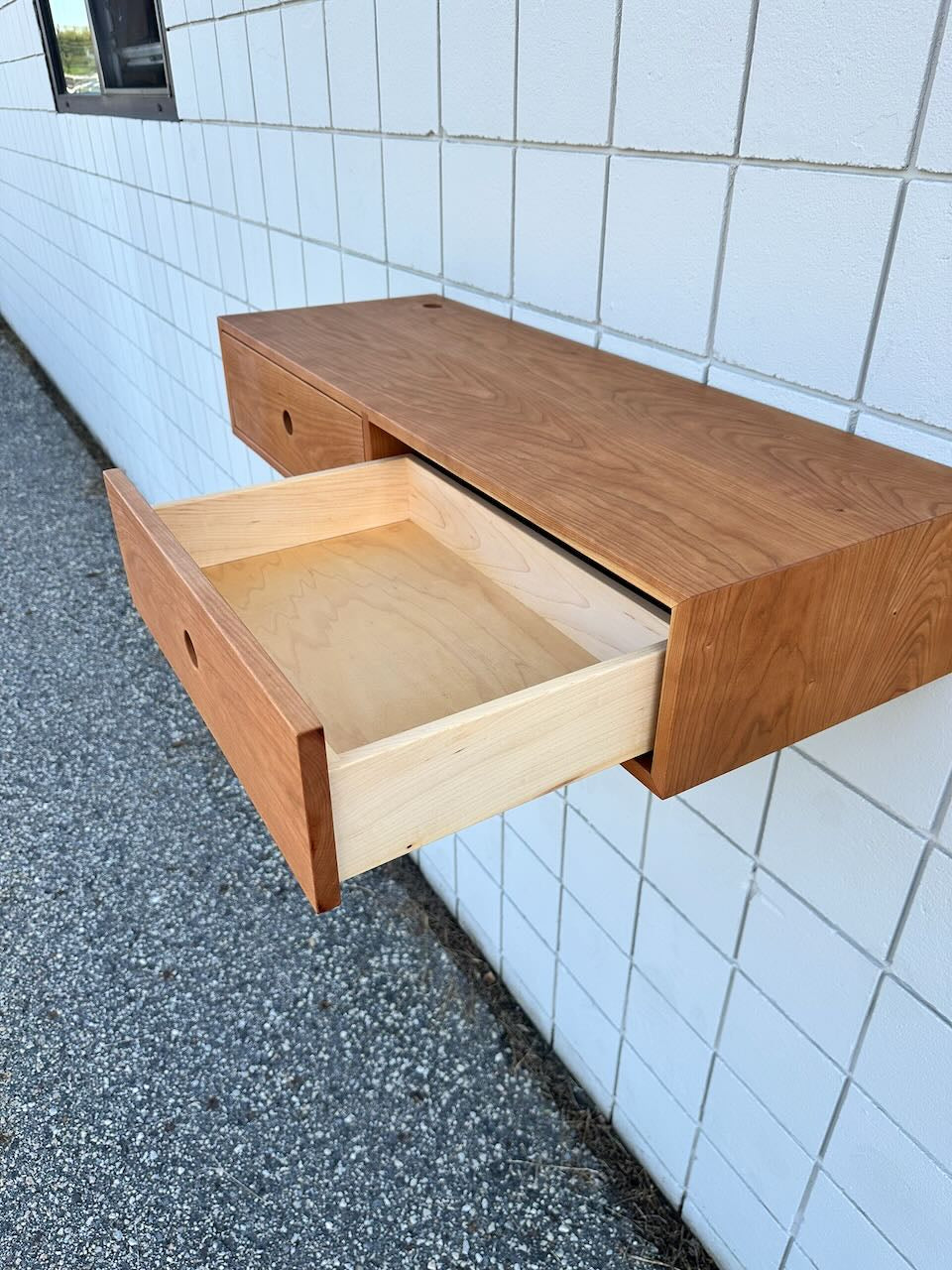 A Krovel Furniture Co. Cherry Desk 40″ - SAMPLE, featuring a floating design with two drawers, is mounted on a white tiled outdoor wall. One solid Maple drawer is open to reveal its light wood interior, while the other stays closed. Gravel covers the ground beneath the unit.