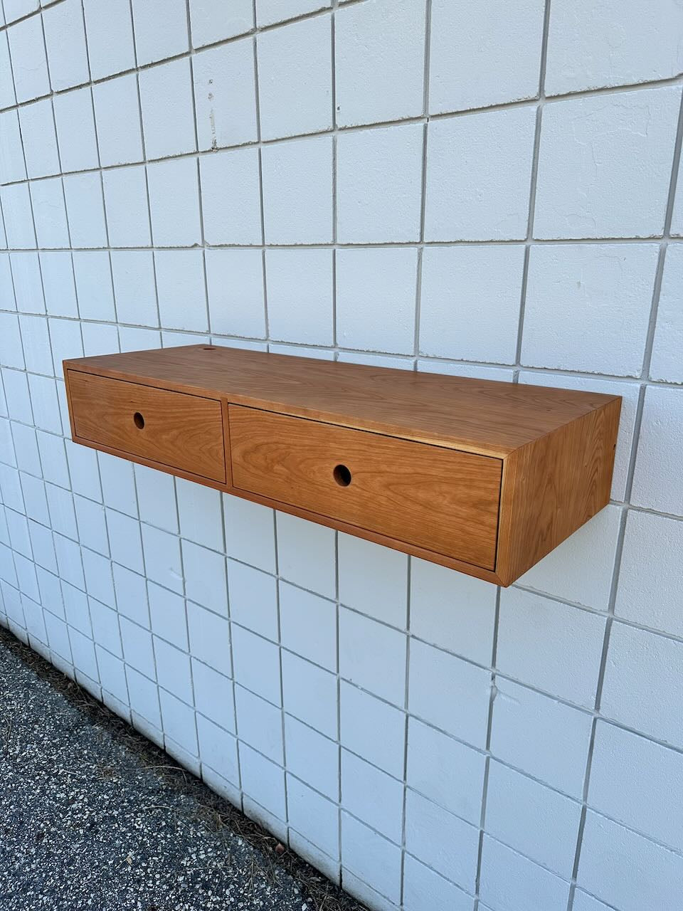A Krovel Furniture Co. Cherry Desk 40″ - SAMPLE is mounted on a white tiled wall above a speckled concrete floor.