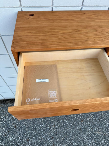 An open wooden drawer, crafted from solid Maple drawers, contains a closed brown folder labeled “wildpac” with a QR code, resting on a light wood interior. The drawer is part of the Cherry Desk 40″ - SAMPLE by Krovel Furniture Co., which is positioned against a tiled wall.