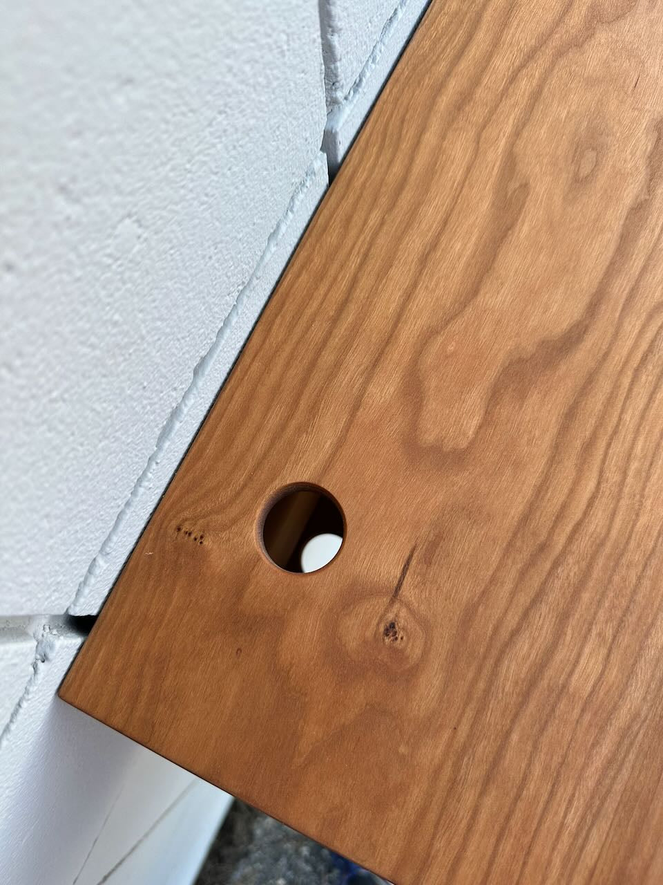 A close-up of the Krovel Furniture Co. Cherry Desk 40″ - SAMPLE shows its wooden surface with a circular hole near the edge, positioned next to a white textured wall with visible brick seams.