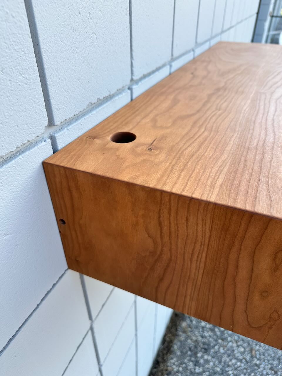 A close-up of a wooden shelf mounted on a white tiled wall, showing a round hole on the top surface near the wall. The design subtly echoes the clean lines and craftsmanship of Krovel Furniture Co.’s Cherry Desk 40″ - SAMPLE, lending an elegant, floating desk aesthetic to the space.