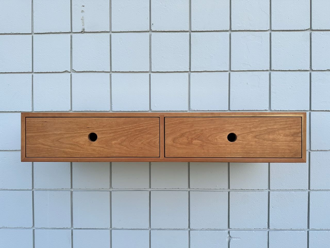 A floating wooden cabinet, the Cherry Desk 40″ by Krovel Furniture Co., features solid Maple drawers with round hole handles and is mounted on a white tiled wall.