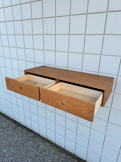 A Cherry Desk 40″ - SAMPLE by Krovel Furniture Co., featuring solid Maple drawers and two open compartments, is mounted on a white tiled wall. Reminiscent of a floating desk, the wooden cabinet hovers elegantly above the concrete floor.