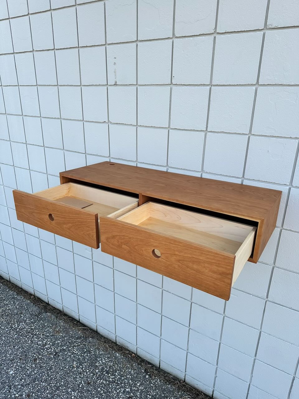 A Cherry Desk 40″ - SAMPLE by Krovel Furniture Co., featuring solid Maple drawers and two open compartments, is mounted on a white tiled wall. Reminiscent of a floating desk, the wooden cabinet hovers elegantly above the concrete floor.
