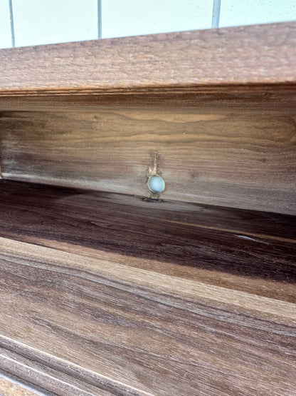 A close-up of a wooden shelf showcases a small white plastic screw cover attached to the back panel, creating a floating effect. The visible wood grain and hint of wall tile evoke the refined style of the Krovel Furniture Co. Custom Walnut Media Table – SECOND.