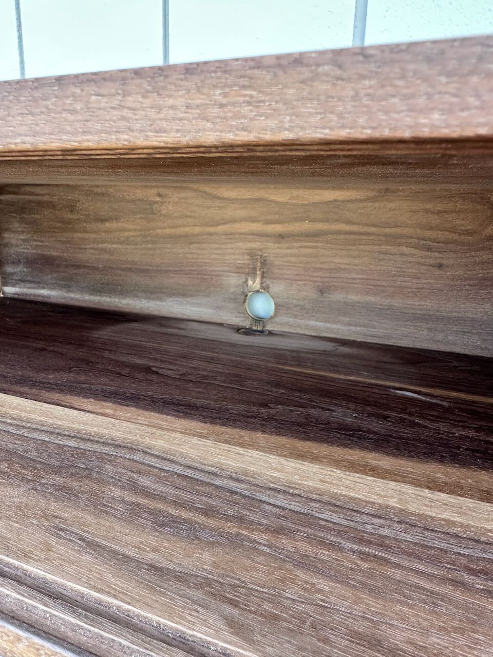 A close-up of a wooden shelf showcases a small white plastic screw cover attached to the back panel, creating a floating effect. The visible wood grain and hint of wall tile evoke the refined style of the Krovel Furniture Co. Custom Walnut Media Table – SECOND.