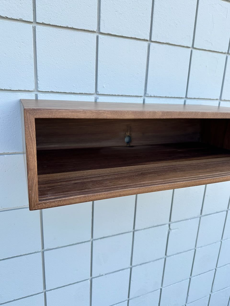 A Custom Walnut Media Table - SECOND by Krovel Furniture Co. is mounted on a tiled white wall, creating a clean floating appearance. The table features an open front design and has a visible screw hole at the back for mounting.