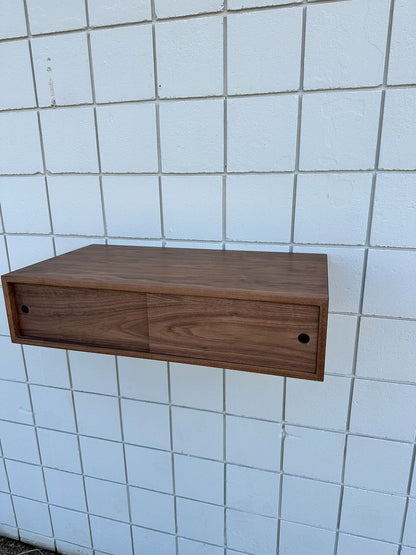 A floating drawer unit by Krovel Furniture Co., inspired by their Custom Walnut Media Table – SECOND, is mounted on a white tiled wall to create a striking floating effect. Crafted with a smooth, natural walnut finish, each of its two drawers features a small, round hole as a handle for minimalist appeal.