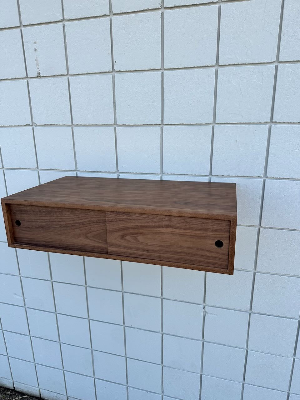 A floating drawer unit by Krovel Furniture Co., inspired by their Custom Walnut Media Table – SECOND, is mounted on a white tiled wall to create a striking floating effect. Crafted with a smooth, natural walnut finish, each of its two drawers features a small, round hole as a handle for minimalist appeal.