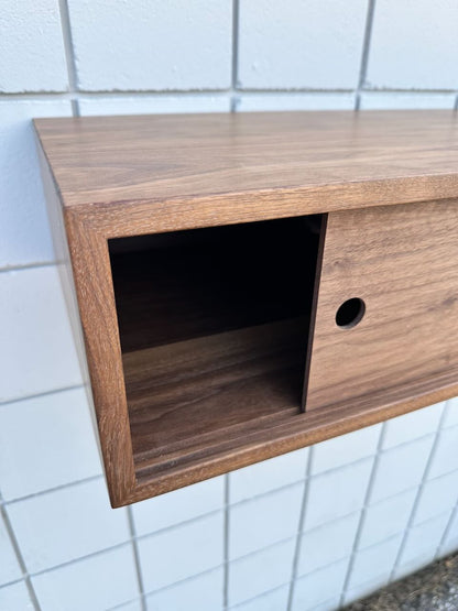 Close-up of a wooden wall shelf with sliding doors, mounted on a white tiled wall. The shelf has a floating look and features a partially open sliding door revealing an empty compartment inside, echoing the sleek design of the Custom Walnut Media Table - SECOND by Krovel Furniture Co.