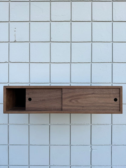 A wall-mounted wooden shelf inspired by the Custom Walnut Media Table - SECOND from Krovel Furniture Co., featuring two sliding doors and an open compartment for a floating effect against a white tiled wall.