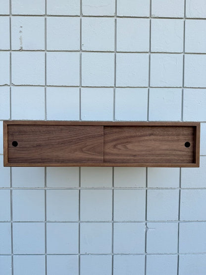 A rectangular wooden wall-mounted cabinet with two front sliding doors, set against a backdrop of white tiles, creates a floating effect reminiscent of the sleek Custom Walnut Media Table – SECOND by Krovel Furniture Co.