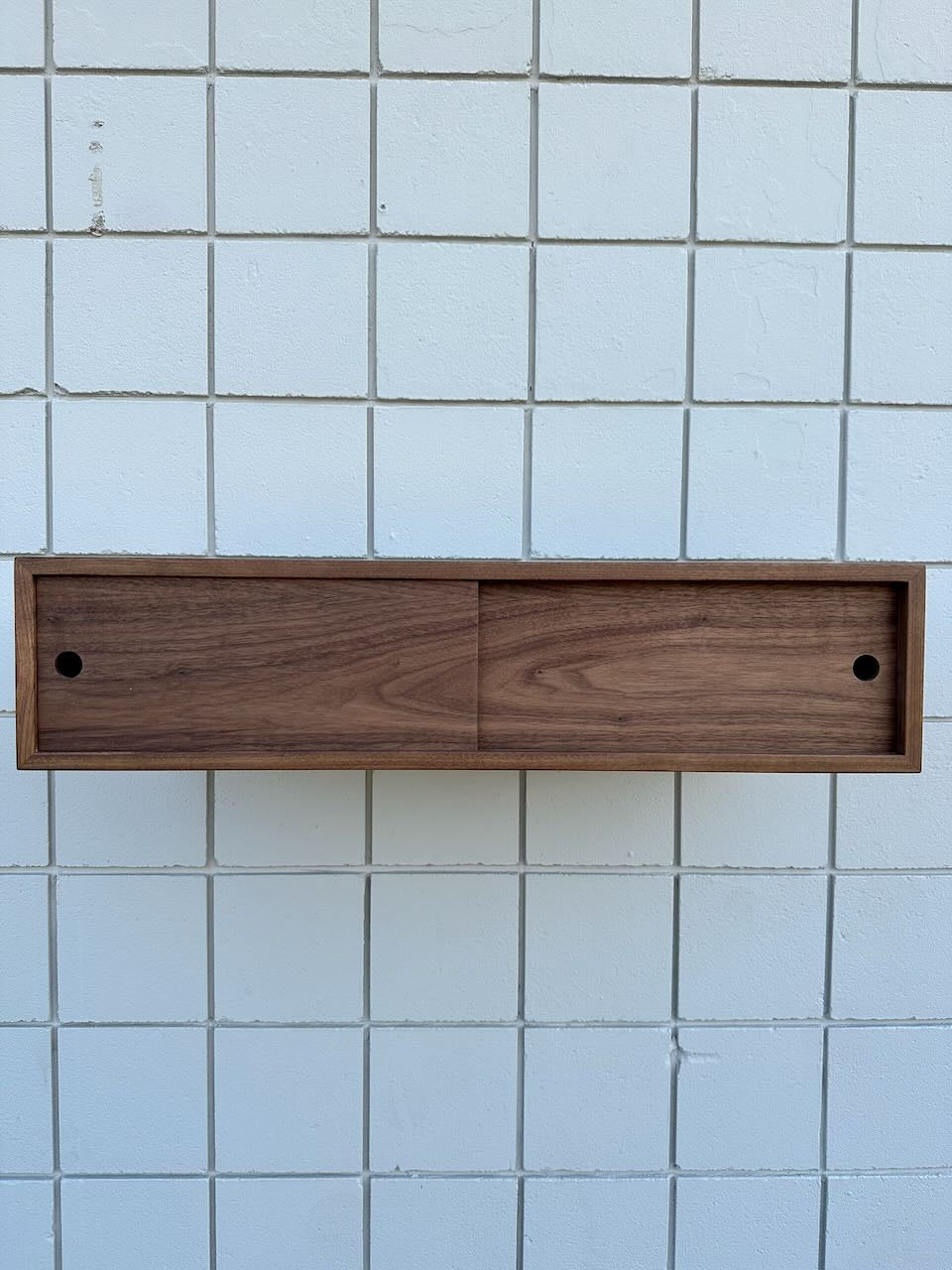 A rectangular wooden wall-mounted cabinet with two front sliding doors, set against a backdrop of white tiles, creates a floating effect reminiscent of the sleek Custom Walnut Media Table – SECOND by Krovel Furniture Co.