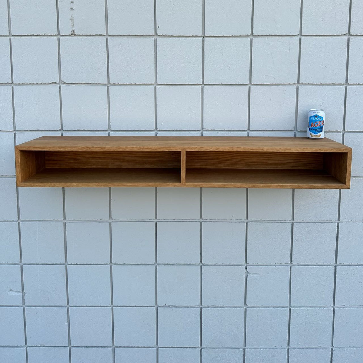 A 48″ White Oak Console Table by Krovel Furniture Co. is mounted against a white tiled wall, its two open compartments reflecting clean, modern lines. On the top right corner of this VOC-free furniture piece, a can of La Croix sparkling water is placed.