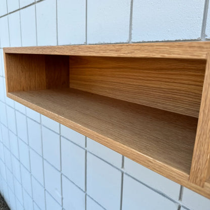 A close-up of the Krovel Furniture Co. 48″ White Oak Console Table - SAMPLE, a rectangular floating shelf mounted on a white tiled wall. The console’s empty, minimalist design highlights its visible white oak grain, showcasing the natural beauty characteristic of VOC-free furniture.