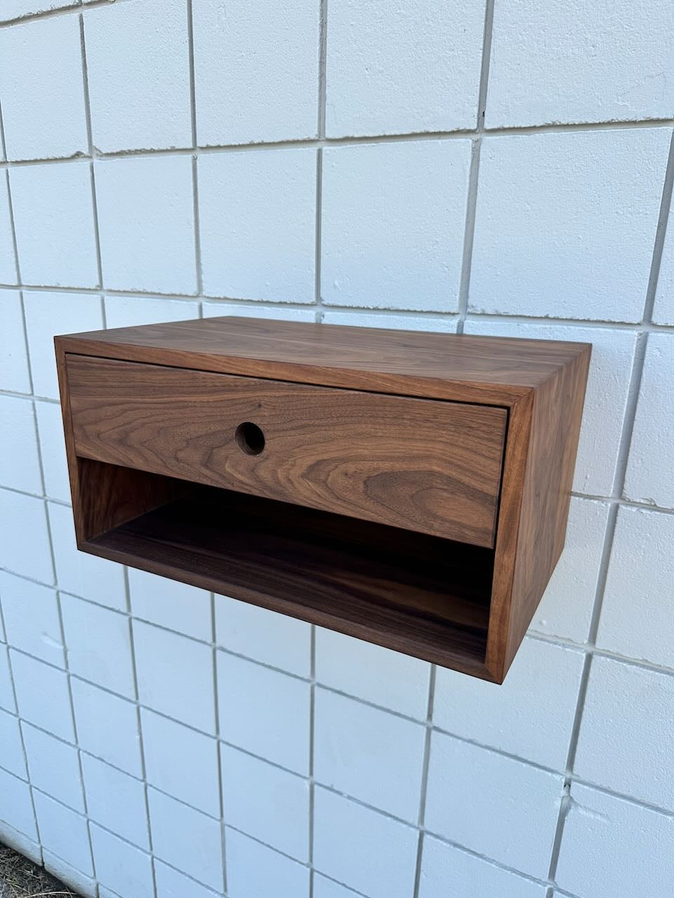 A pair of walnut floating nightstands by Krovel Furniture Co., each featuring solid Hard Maple drawers, an open storage space below, and a VOC-free finish, are mounted on a white tiled wall.