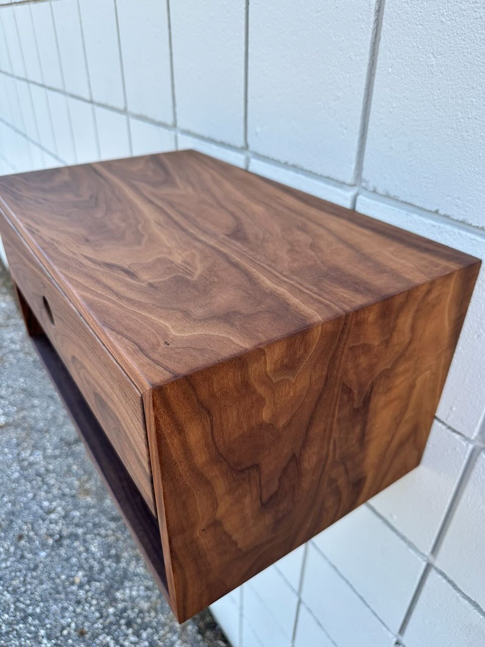 A PAIR (2) of Walnut Floating Nightstands by Krovel Furniture Co. is mounted on a white tiled wall. These nightstands feature a rich brown wood grain finish with sharp, clean edges, and are expertly crafted in the signature Krovel style. Each includes a solid Hard Maple drawer for durability, and the VOC-free finish ensures an eco-friendly touch.