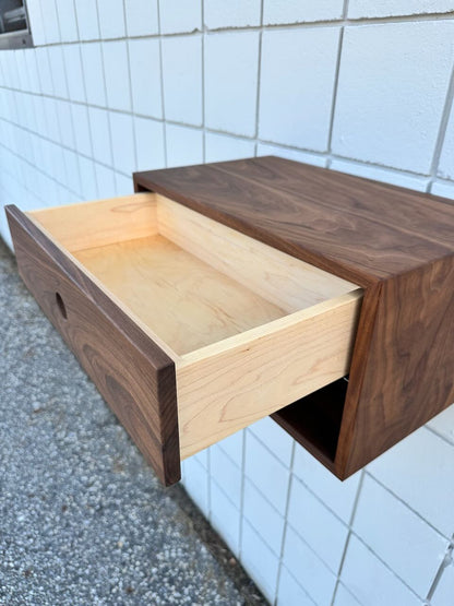 Here’s a rewritten version of your sentence using the provided product data: A PAIR (2) of Walnut Floating Nightstands by Krovel Furniture Co. are mounted on a white tiled wall, each featuring a dark brown walnut exterior and drawers crafted from solid Hard Maple with a light wood interior. One nightstand is pulled open to reveal its empty inside.
