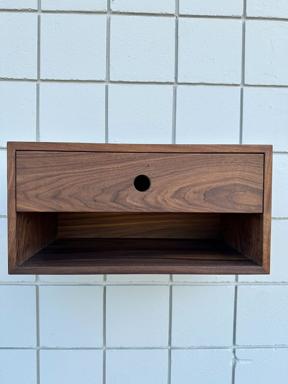 A PAIR (2) of walnut floating nightstands by Krovel Furniture Co.—sample pieces that channel the elegance of minimalist wall-mounted design—each features a solid Hard Maple drawer with a circular cutout handle, set against a white tiled wall, and finished with a VOC-free coating for a safer home environment.