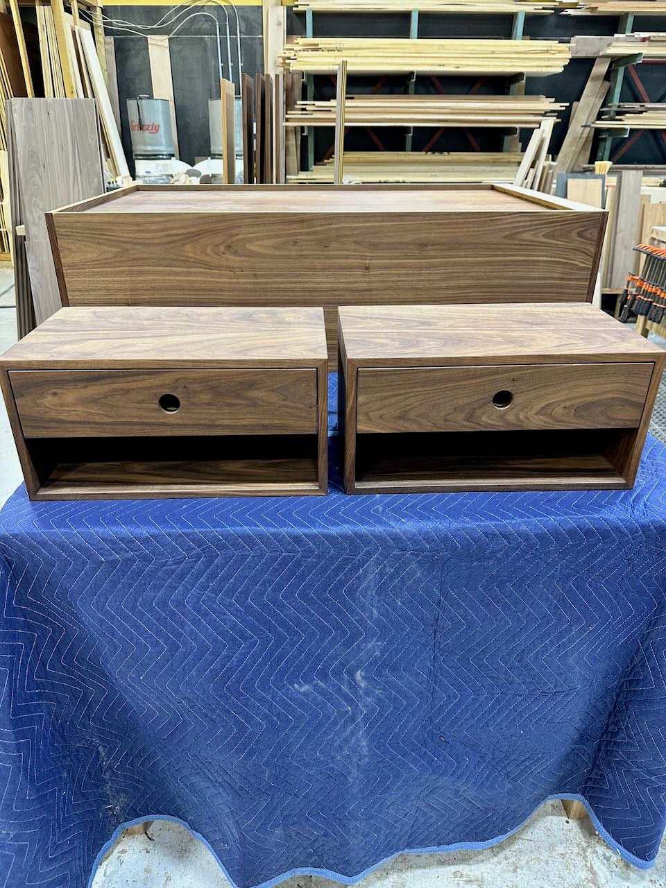 Two small walnut floating nightstands from Krovel Furniture Co. (PAIR (2) Walnut Floating Nightstands - SAMPLES) and one larger cabinet are displayed on a blue fabric in a workshop with wood panels and tools in the background.