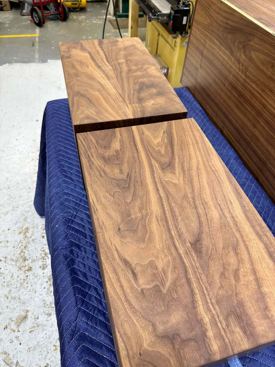 Two walnut boards with rich, natural grain patterns rest on a blue padded surface in a workshop. The boards are aligned, similar to the meticulous craftsmanship seen in Krovel Furniture Co.’s PAIR (2) Walnut Floating Nightstands - SAMPLES. Woodworking tools and surfaces appear in the background.