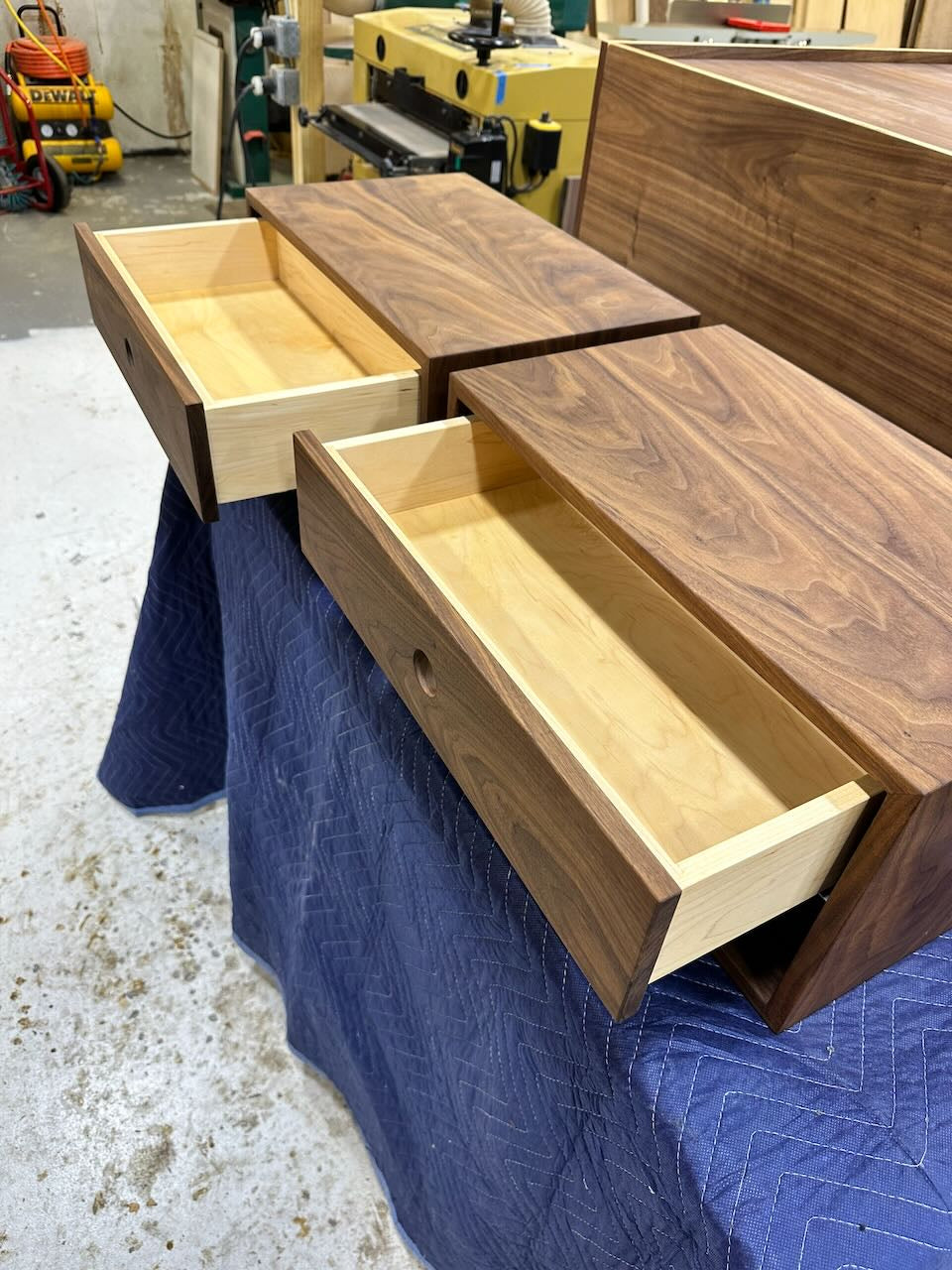 A pair of wooden drawers with dovetail joints, crafted from solid Hard Maple and partially pulled out from a matching cabinet, are displayed on a blue padded cover in a workshop setting. These drawers are featured as samples for the PAIR (2) Walnut Floating Nightstands by Krovel Furniture Co. and come with a durable, VOC-free finish.