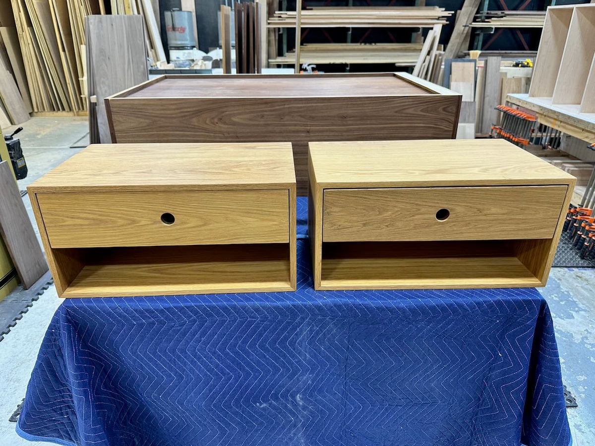 A PAIR (2) of White Oak Floating Nightstands - SECONDS by Krovel Furniture Co. are displayed on a blue padded blanket in a workshop, featuring a single drawer and open shelf, with a larger piece of furniture and various wood materials visible in the background.