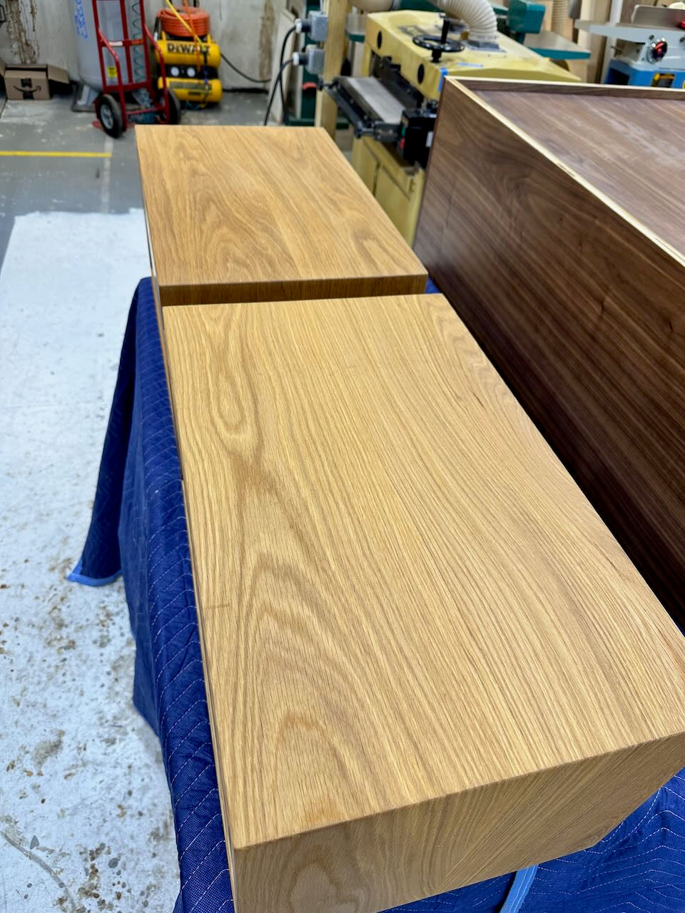 A PAIR (2) of White Oak Floating Nightstands - SECONDS by Krovel Furniture Co. are displayed on a blue quilted blanket in a workshop, surrounded by woodworking tools and machinery, showcasing their solid white oak construction and natural grain finish.