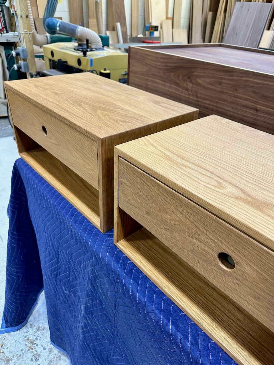 A PAIR (2) of White Oak Floating Nightstands - SECONDS by Krovel Furniture Co., each with open shelves and single drawers featuring round hole drawer pulls, rest on a blue padded surface in a woodworking shop.