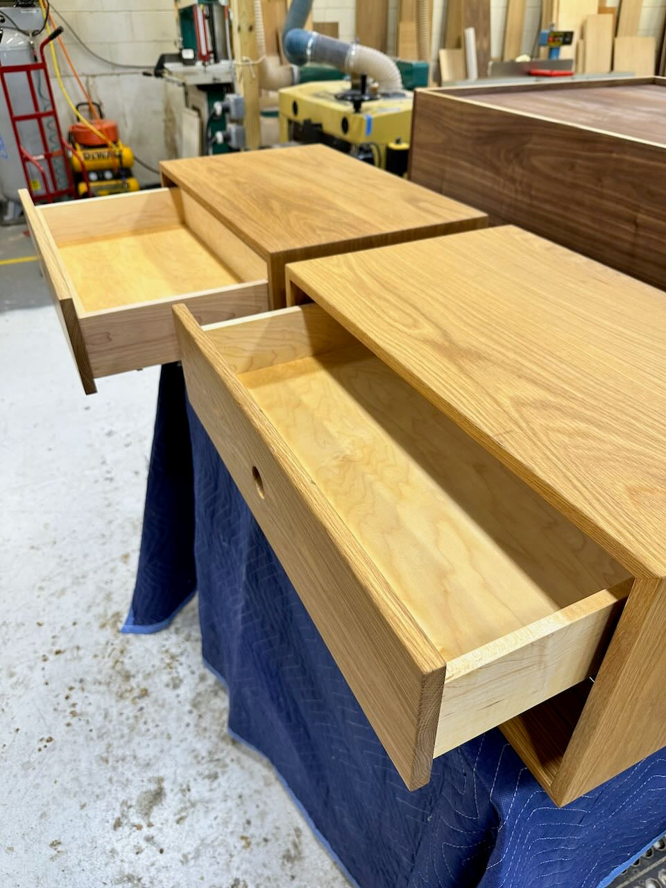 Two wooden drawers are open, revealing their light-colored interiors. The drawers belong to the PAIR (2) White Oak Floating Nightstands - SECONDS by Krovel Furniture Co., shown in a workshop with woodworking tools and equipment in the background.