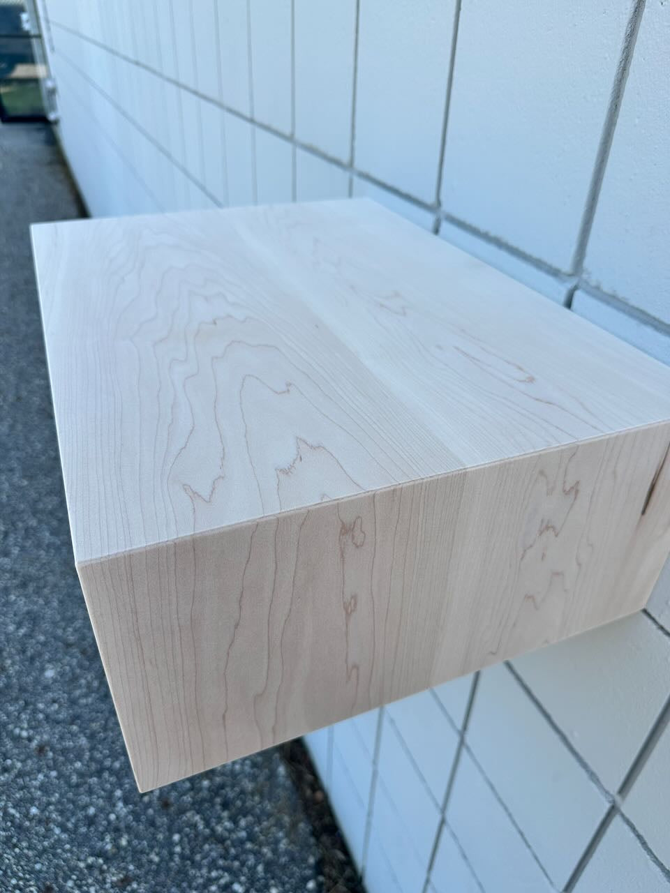 A light-colored wooden shelf with visible natural wood grain, photographed outdoors against a white tiled wall.