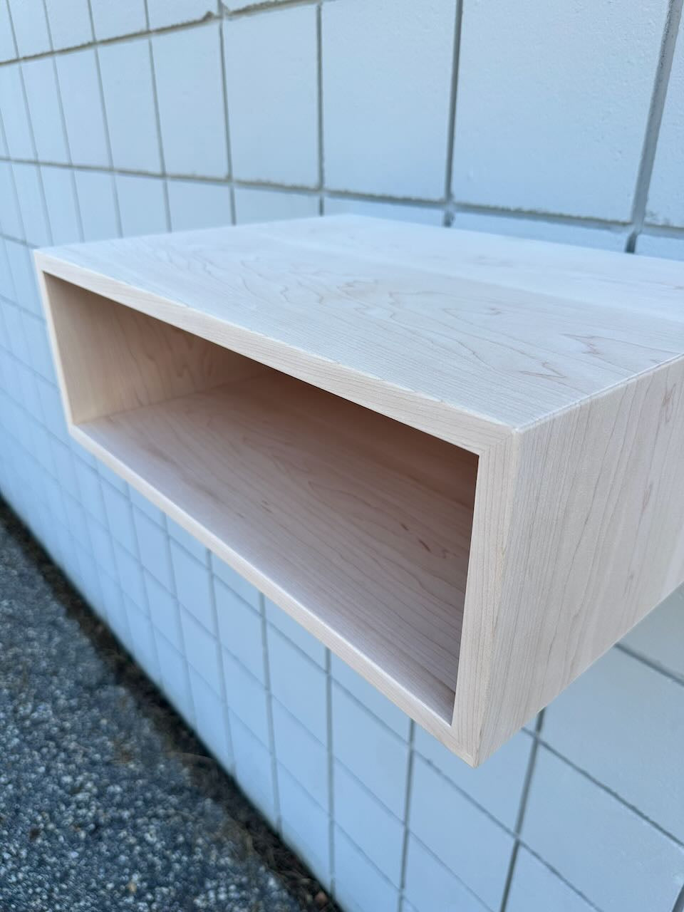 A light natural wood floating wall shelf mounted on a white tiled exterior wall above a gravel ground.