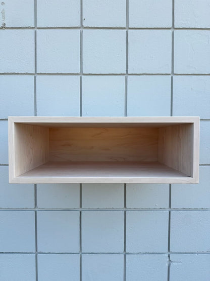 A minimalist, rectangular wooden floating shelf with a light natural finish, mounted on a light blue tiled wall.